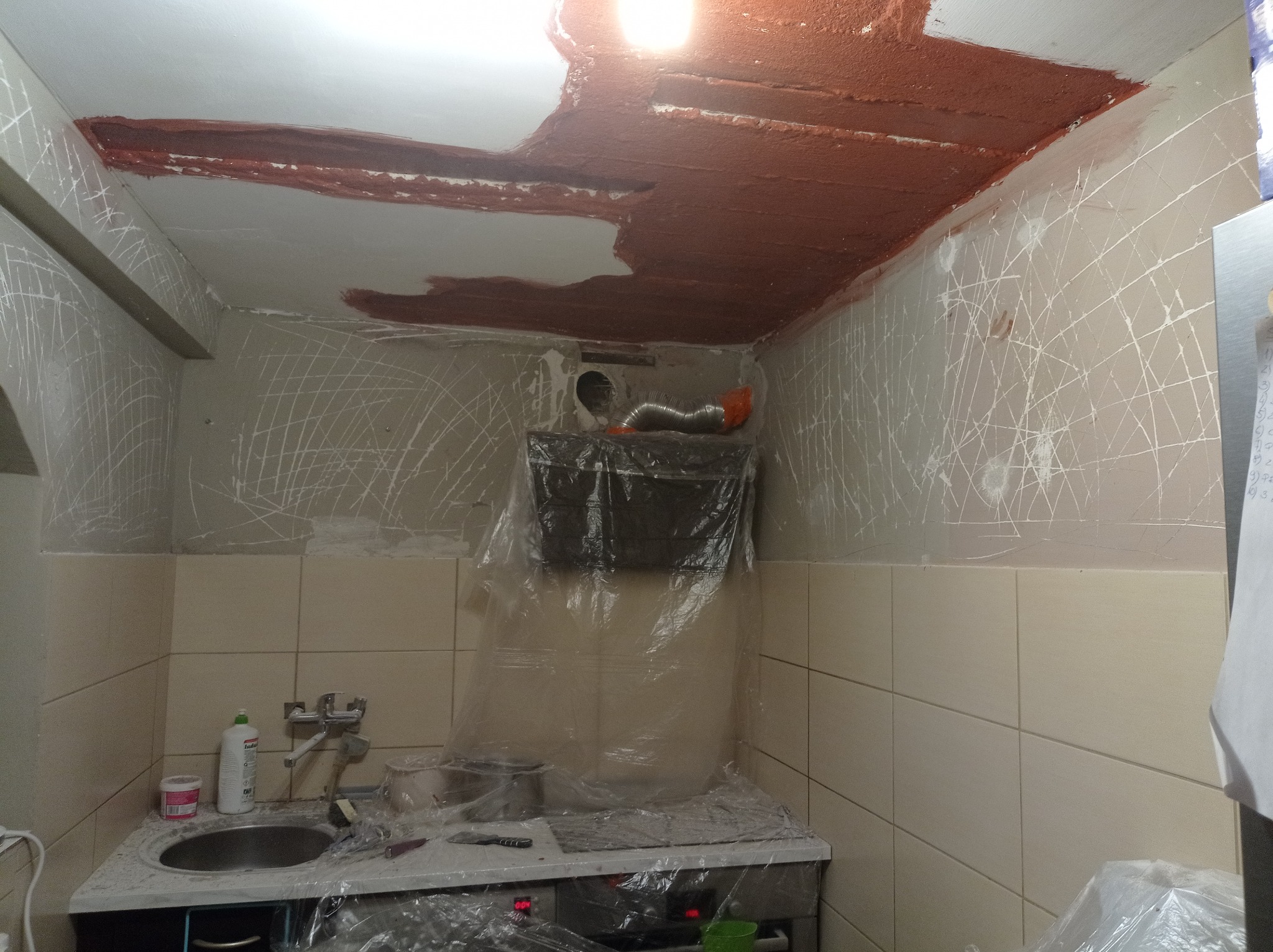 Kuchnia w trakcie przygotowania do prac wykończeniowych: ściany zarysowane pod tynk, sufit częściowo pokryty brązową warstwą, zabezpieczone blaty i sprzęty folią.