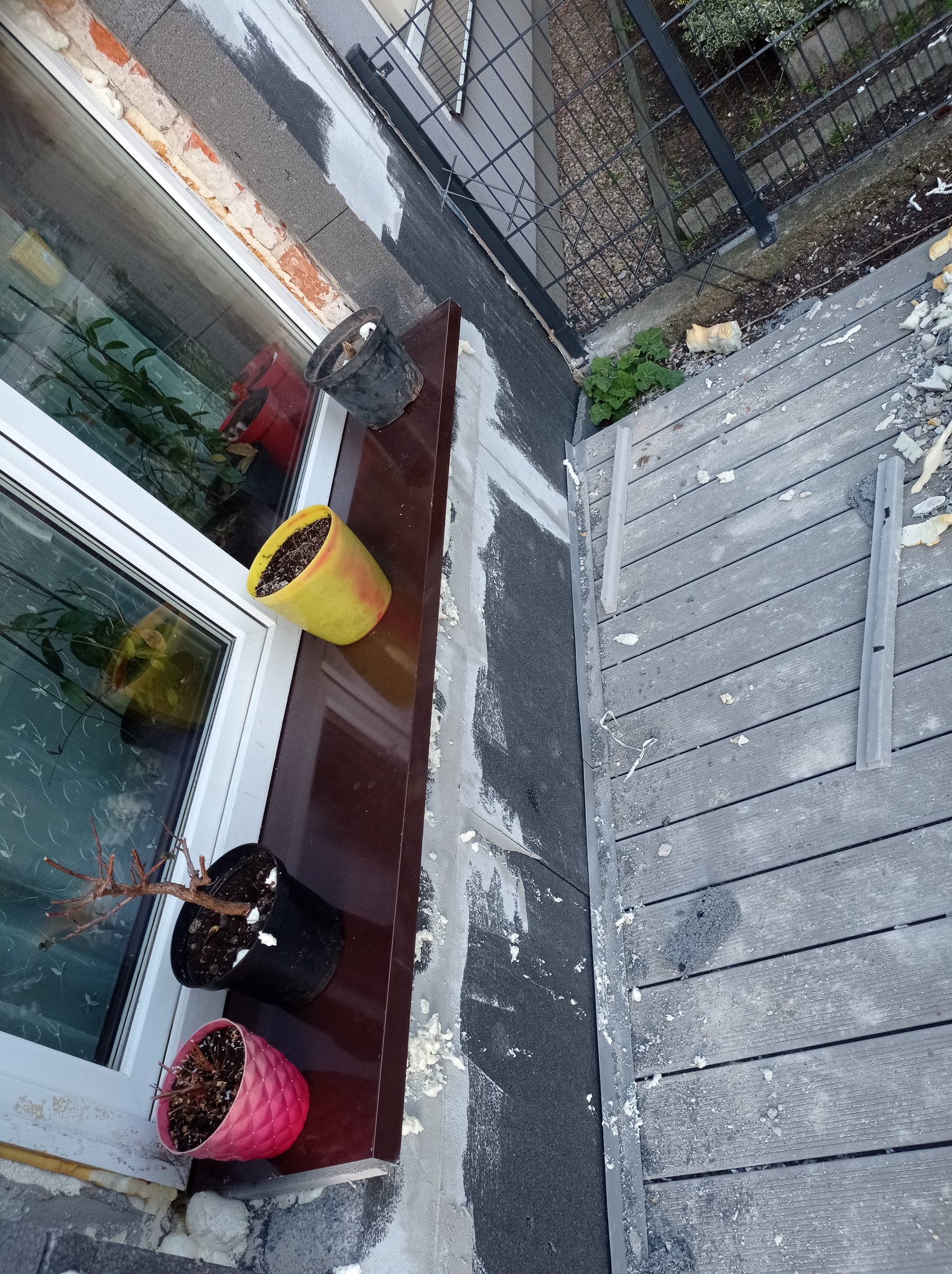 Widok z góry na okno z doniczkami podczas prac wykończeniowych elewacji, z widoczną pianką montażową i deskami tarasowymi na zewnątrz.