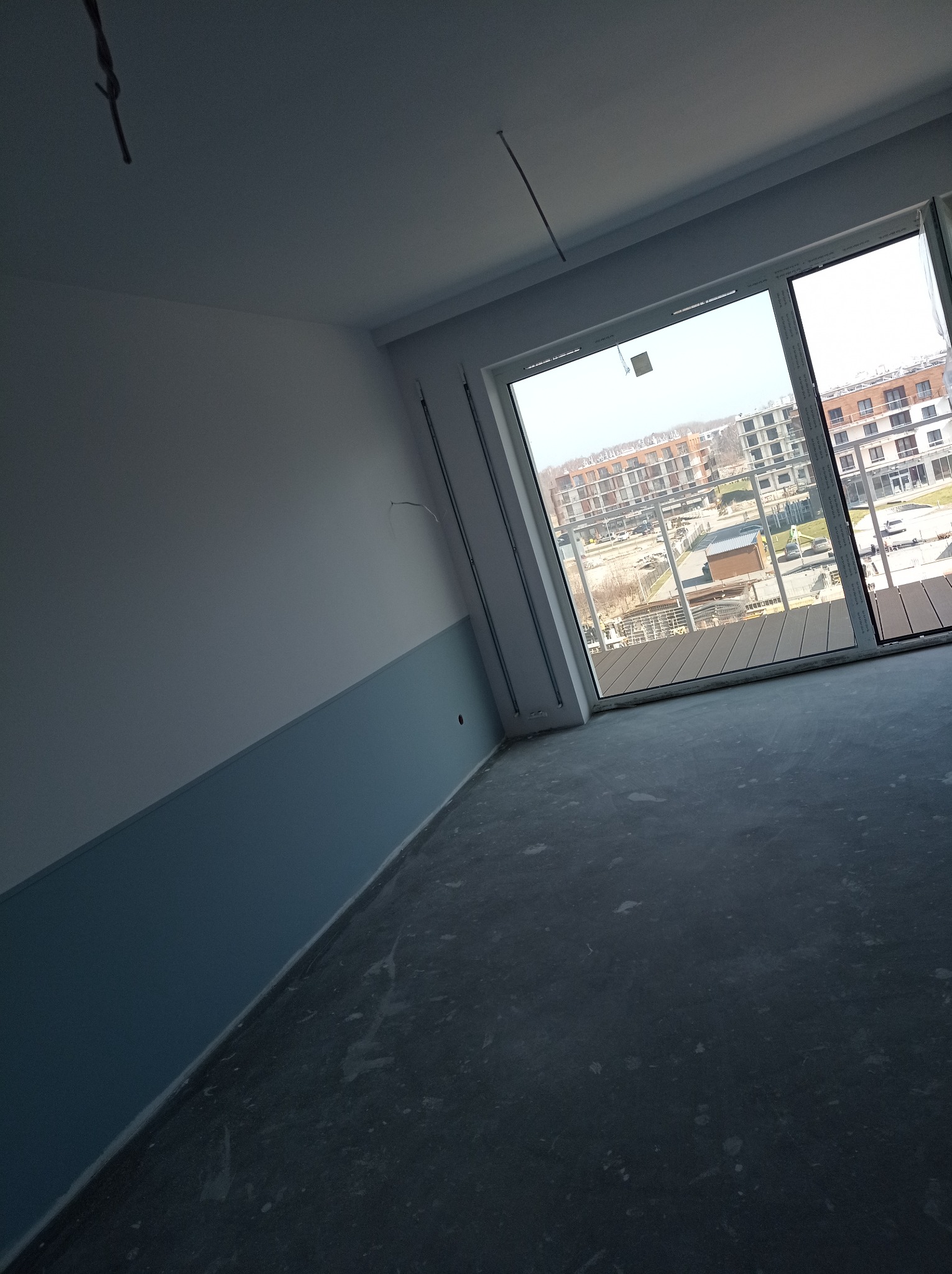 Wnętrze w trakcie prac wykończeniowych: ściana częściowo pomalowana na niebiesko, surowa podłoga, widok na osiedle z balkonu przez duże okno.
