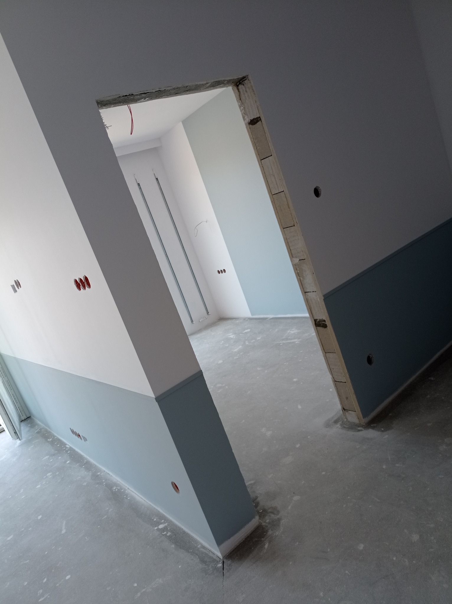 Surowe wnętrze w trakcie prac wykończeniowych: pomalowane ściany na biało i niebiesko, widoczne otwory pod gniazdka elektryczne i surowa podłoga, z widocznym otworem drzwiowym bez ościeżnicy.