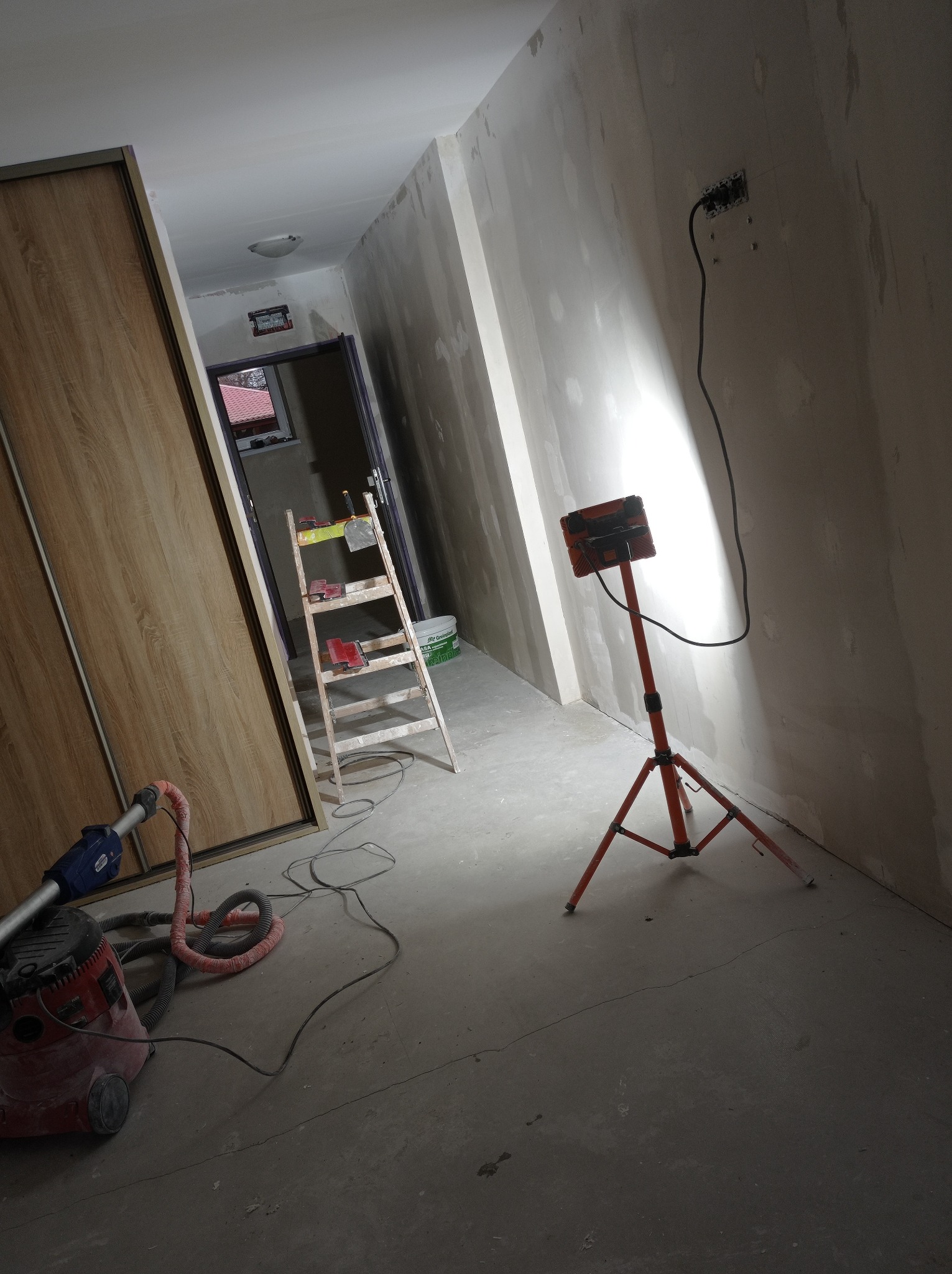 Wnętrze w trakcie prac przygotowawczych: ściany po szpachlowaniu, drabina z narzędziami, lampa oświetleniowa na trójnogu, odkurzacz przemysłowy przy szafie wnękowej.