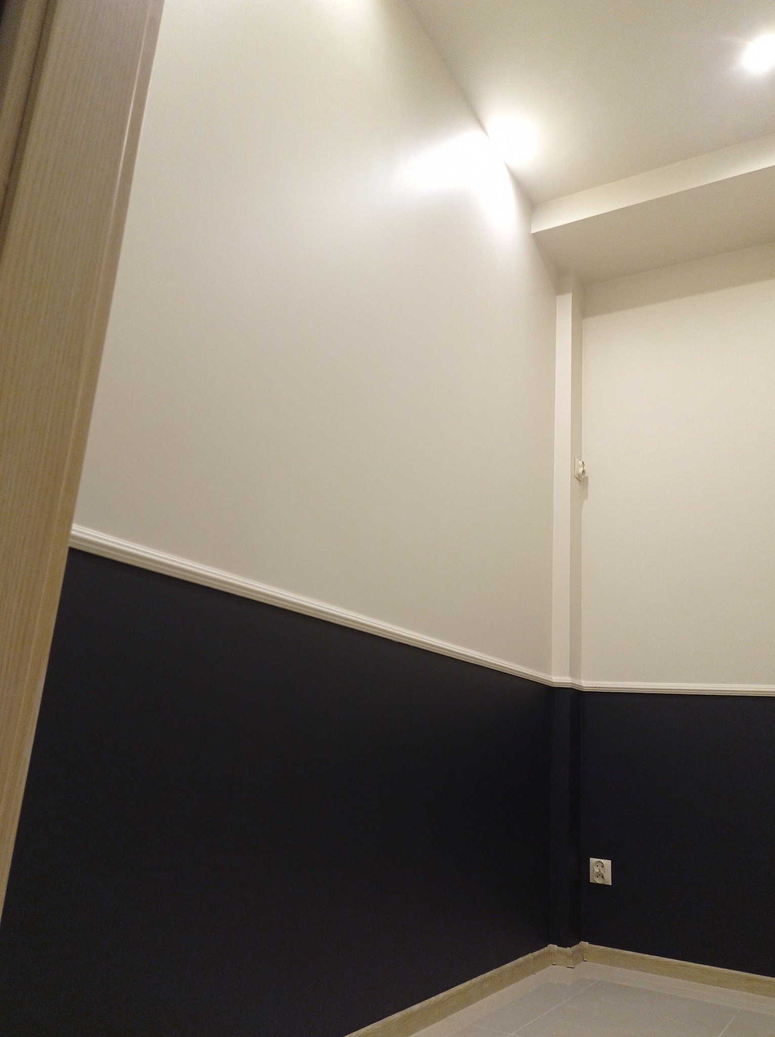 Świeżo pomalowane wnętrze z dwukolorową ścianą (biała góra, granatowy dół) oddzieloną ozdobną listwą, widoczny fragment drzwi i gniazdko elektryczne.