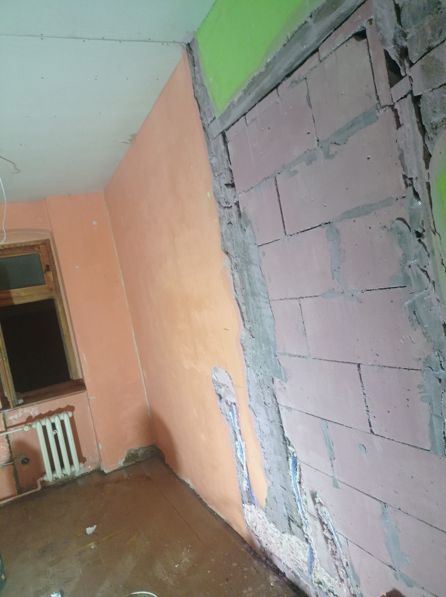 Fragment ściany w trakcie prac przygotowawczych, widoczne odsłonięte bloczki, częściowo nałożona warstwa tynku w odcieniu brzoskwiniowym, stara stolarka okienna i grzejnik żeberkowy w tle.