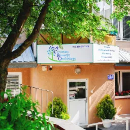 Budynek Centrum Rozwoju Osobistego z zielonym logo drzewa na szyldzie, numer 74A na ścianie, drzewa w donicach i kwiaty w oknie, widoczne zza drzewa.