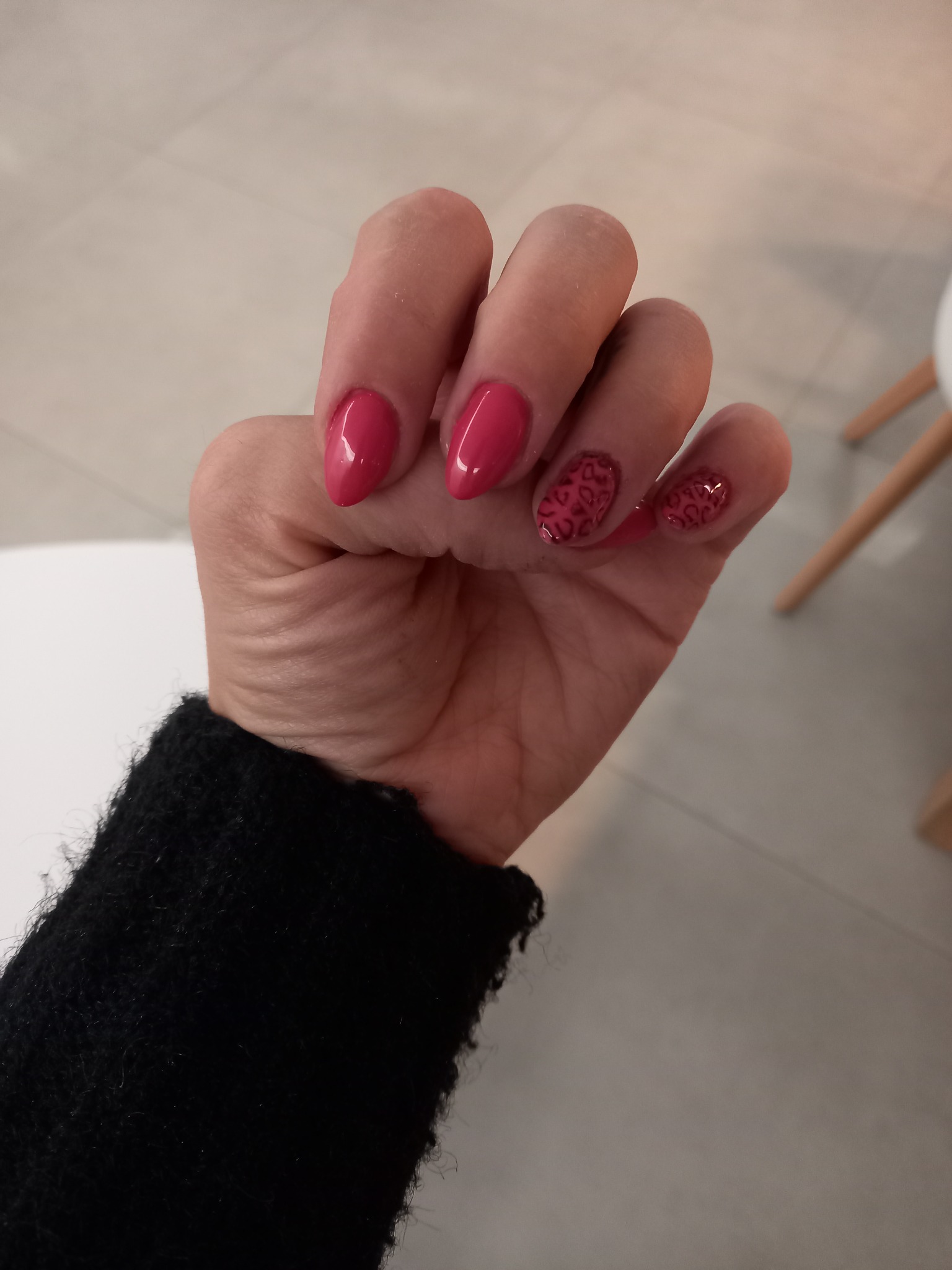 Dłoń z pomalowanymi na różowo paznokciami w kształcie migdała, dwa z nich zdobione wzorem w panterkę, na tle jasnej podłogi i fragmentu czarnego swetra.