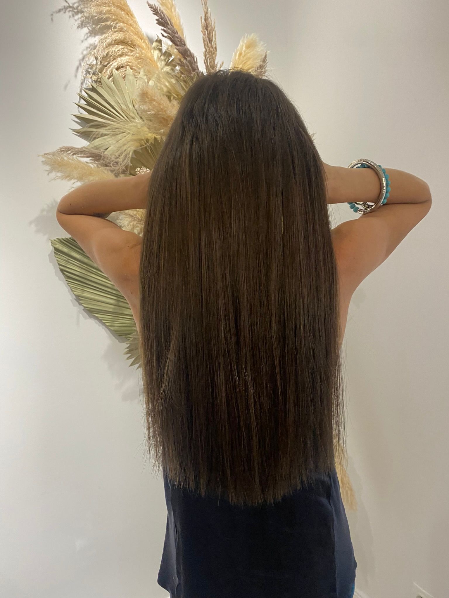 Długie, proste, brązowe włosy kobiety, tyłem do kamery, z rękami uniesionymi do góry, eksponującymi turkusowe bransoletki, na tle białej ściany z dekoracją z suszonych traw.