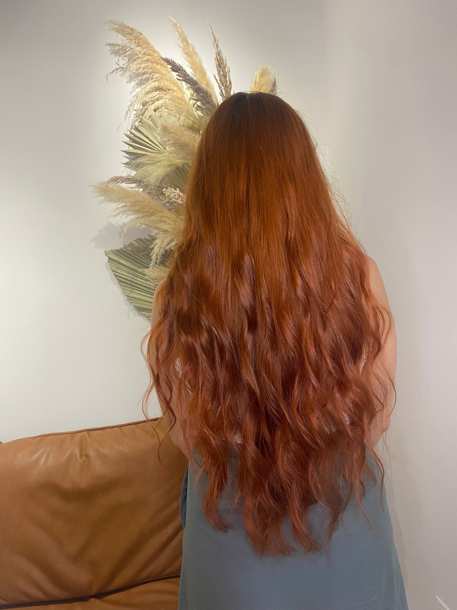 Długie, falowane włosy w odcieniach miedzi, widok z tyłu, dekoracja z suszonych traw w tle, skórzana sofa w kadrze.