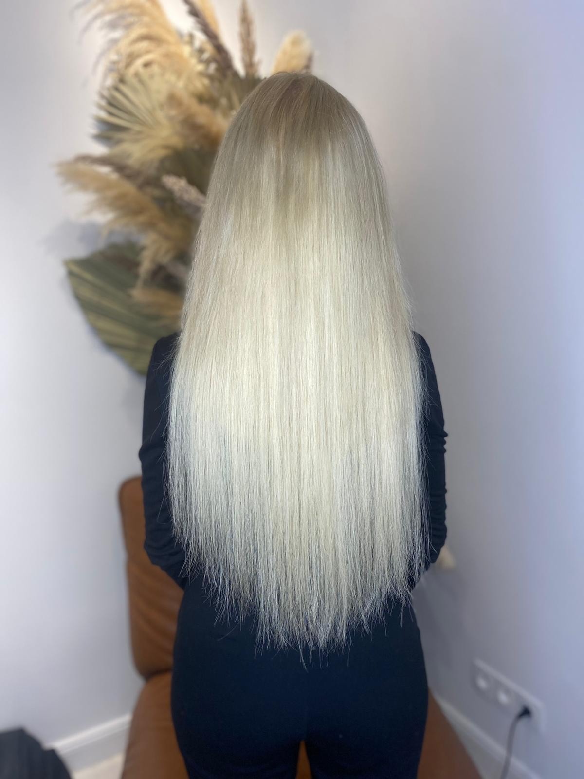 Długie, platynowe blond włosy, widok z tyłu, delikatne cieniowanie u dołu, na tle jasnej ściany z dekoracyjną kompozycją z suszonych traw.