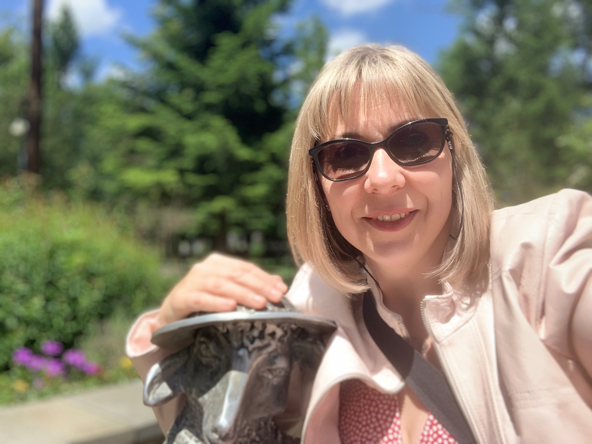 Uśmiechnięta blondynka w okularach przeciwsłonecznych, w różowej kurtce, robiąca selfie na tle zieleni, trzymająca rękę na metalowej rzeźbie głowy zwierzęcia.
