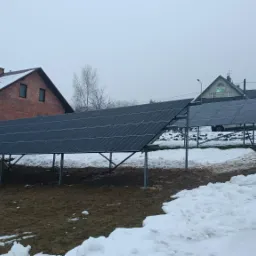 Instalacja fotowoltaiczna na metalowej konstrukcji, częściowo zasypana śniegiem, na tle domów w pochmurny dzień.