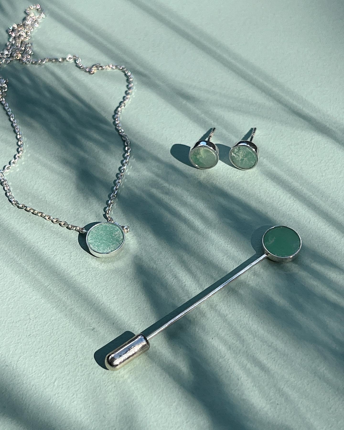 Srebrny naszyjnik z okrągłym, zielonym kamieniem, para kolczyków sztyftów z zielonymi kamieniami i srebrna broszka z zielonym kamieniem na tle turkusowego stołu z cieniami liści.