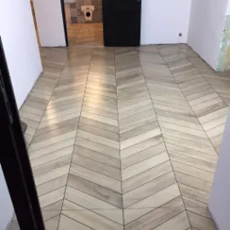 Ułożona pod kątem jasna podłoga z paneli imitujących drewno, widok na otwarte drzwi do łazienki z patchworkowymi kaflami.