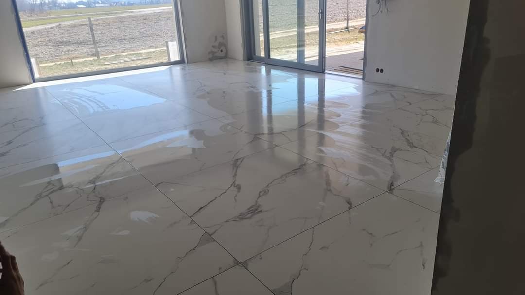 Błyszcząca, biała podłoga z płytek imitujących marmur w nowym, jasnym pomieszczeniu z dużymi oknami; widoczne odbicia światła na powierzchni.