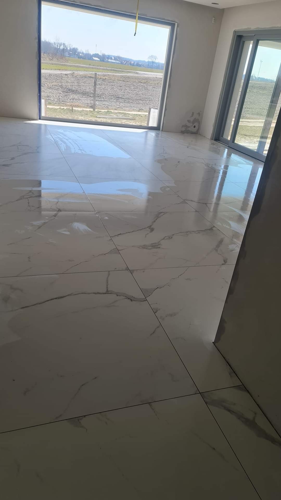 Błyszcząca, marmurowa podłoga z dużych płytek w jasnym, surowym wnętrzu z dużymi oknami wychodzącymi na krajobraz.