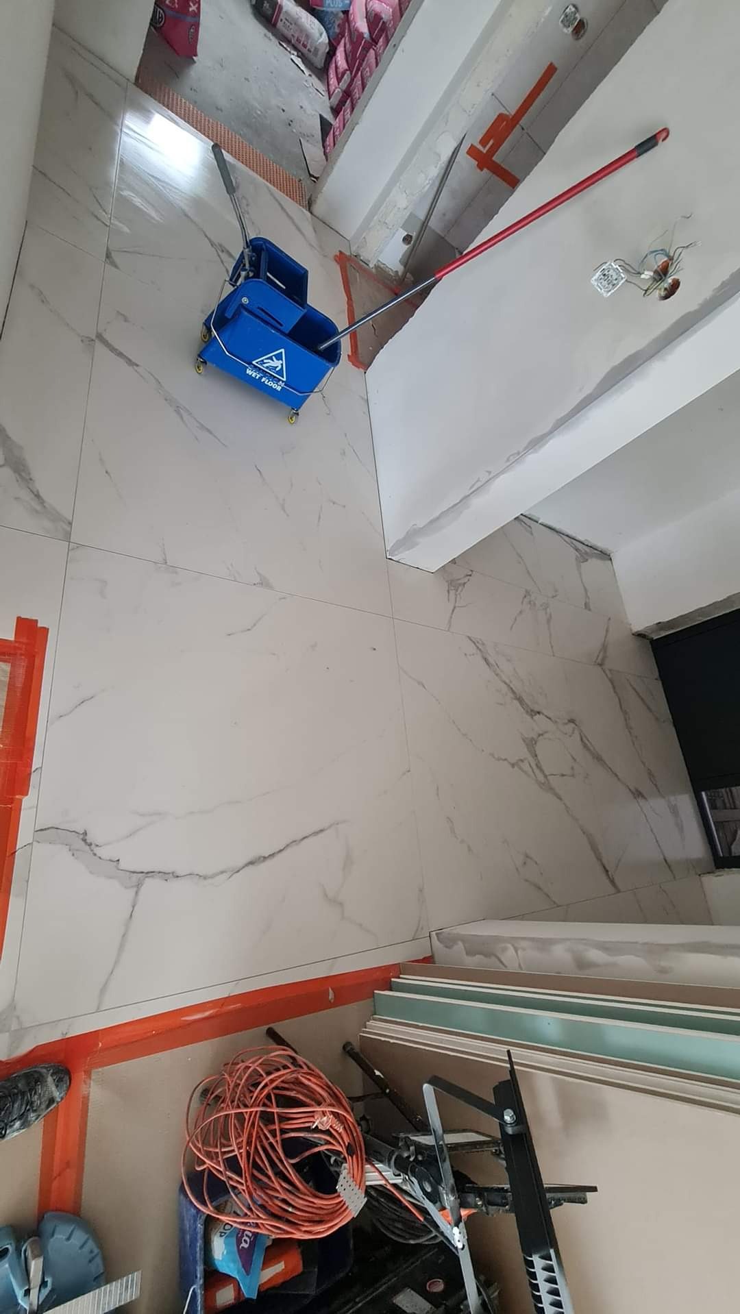 Podłoga wyłożona jasnymi płytkami imitującymi marmur, z widocznym niebieskim wiadrem z mopem na kółkach, pomarańczowym kablem i narzędziami w rogu pomieszczenia, oraz fragmentem ściany z widocznymi...