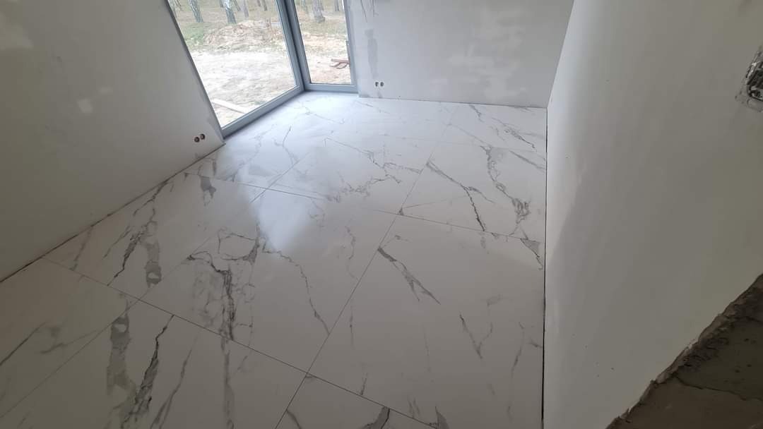 Nowoczesne, białe płytki podłogowe z szarym marmurkowym wzorem, ułożone w jasnym, surowym wnętrzu z dużym oknem i surowymi ścianami.