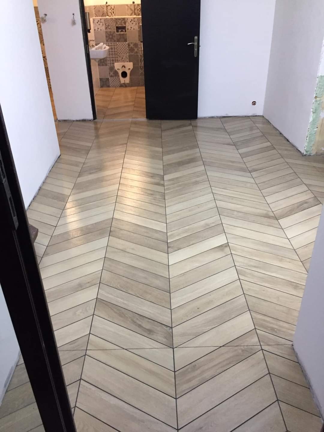 Ułożona pod kątem jasna podłoga z paneli imitujących drewno, widok na otwarte drzwi do łazienki z patchworkowymi kaflami.