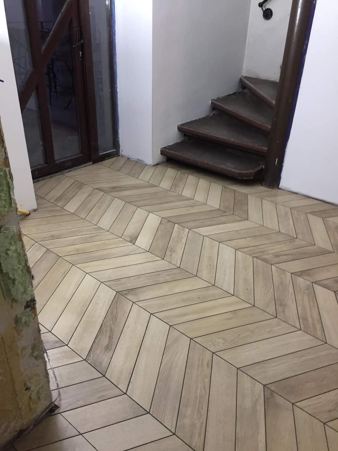 Ułożona podłoga z płytek imitujących drewno, wzór jodełki, widoczne schody i drzwi wejściowe, fragment ściany przed remontem.