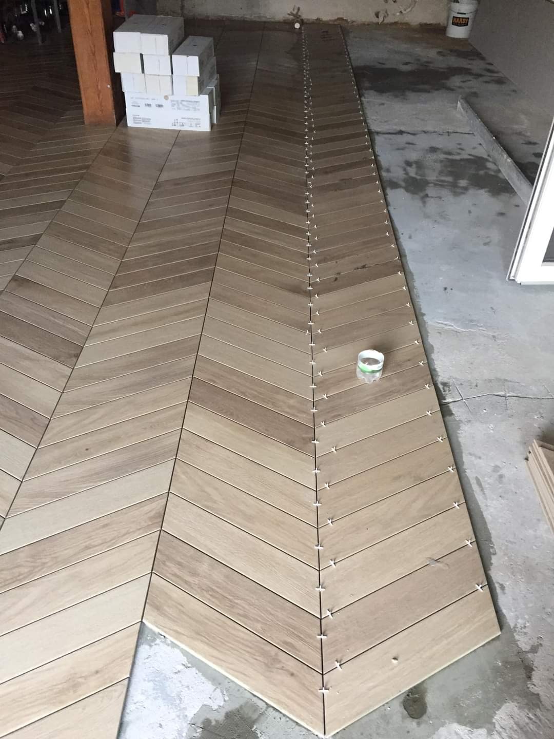Układanie podłogi z płytek imitujących drewno w jodełkę, widoczne krzyżyki dystansowe i zapasowe płytki w kartonach.