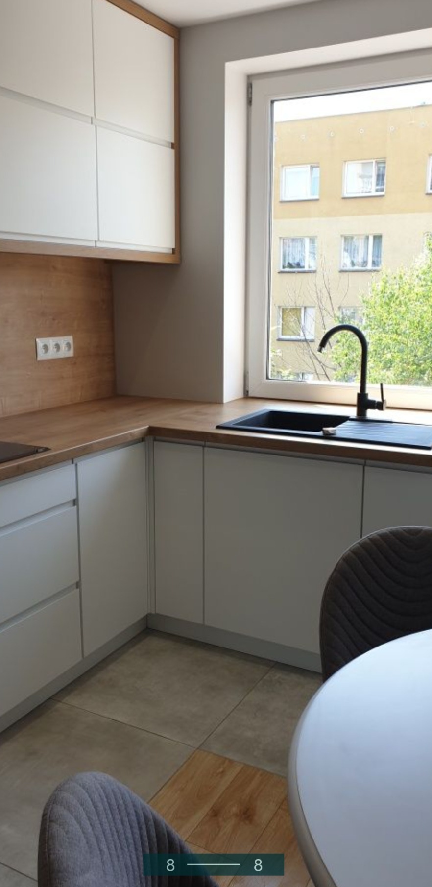 Nowoczesna kuchnia z białymi szafkami, drewnianym blatem i czarną armaturą, widok na budynek przez okno.