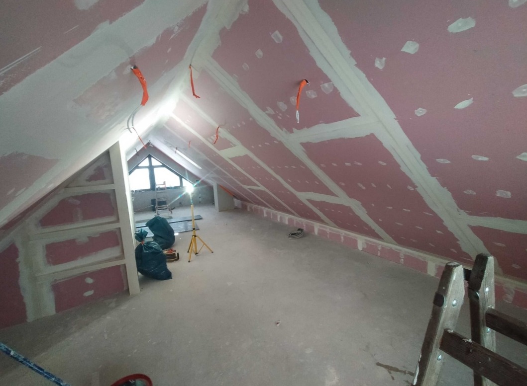 Poddasze w trakcie zabudowy płytami gipsowo-kartonowymi, widoczne szpachlowane łączenia, wystające kable elektryczne i drabina oparta o ścianę.