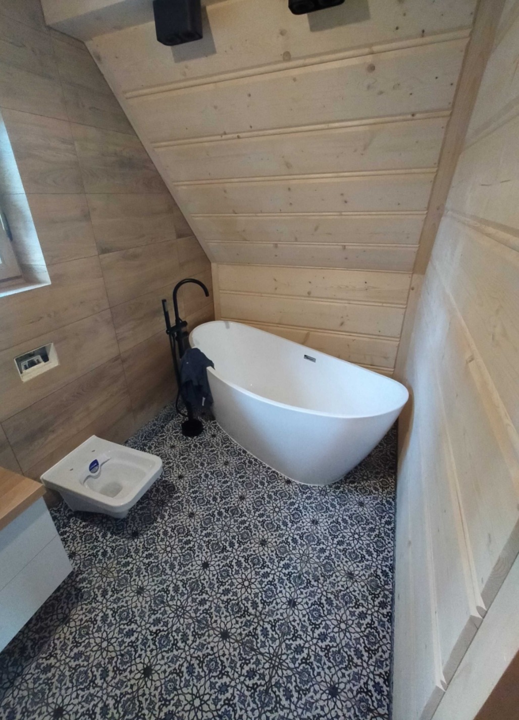 Nowoczesna łazienka na poddaszu z wolnostojącą białą wanną, czarną armaturą, umywalką i wzorzystą podłogą w odcieniach niebieskiego i szarego, wykończona drewnem.