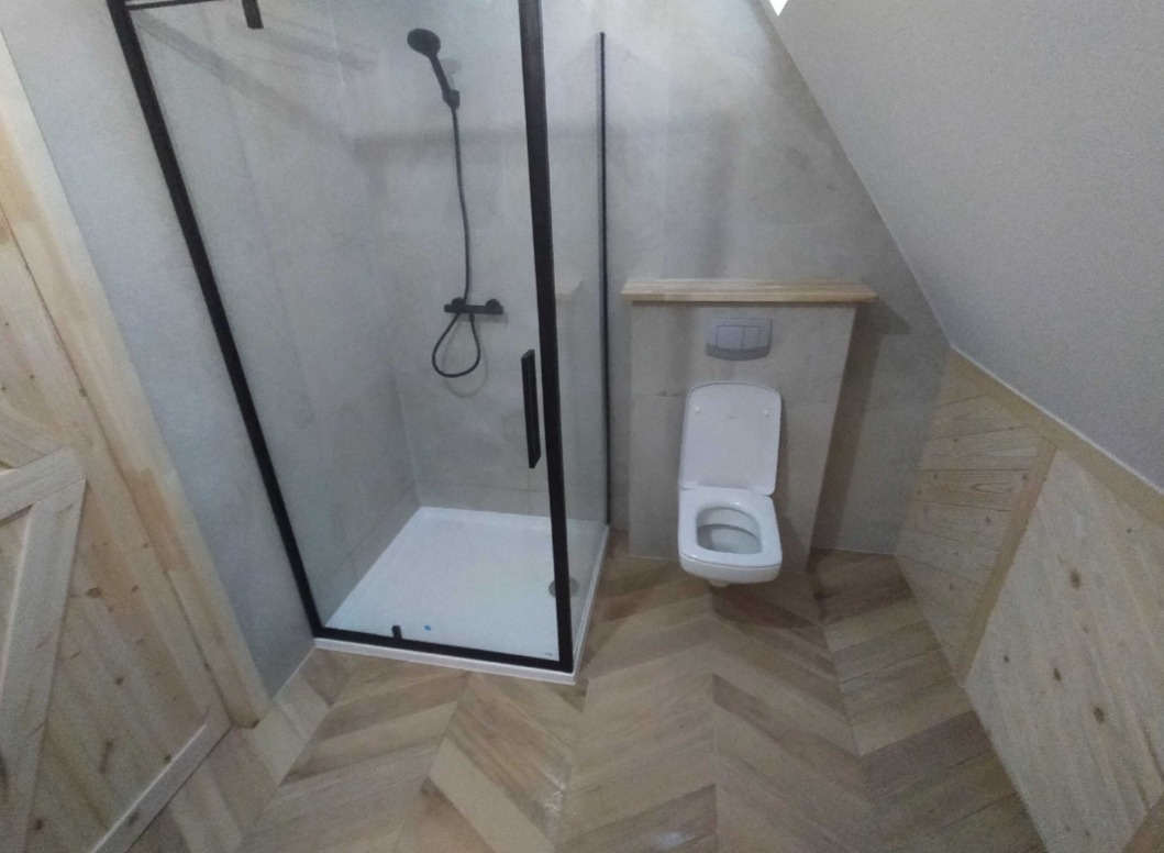 Nowoczesna łazienka na poddaszu z kabiną prysznicową z czarną ramą, toaletą podwieszaną i podłogą ułożoną w jodełkę z jasnego drewna.