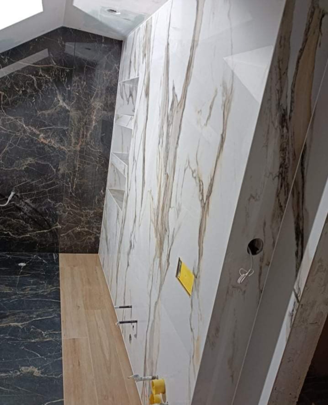 Wnętrze łazienki w trakcie prac wykończeniowych: ściany wyłożone płytkami imitującymi marmur w odcieniach bieli i brązu, z wbudowanymi półkami; podłoga z paneli imitujących drewno; widoczne...