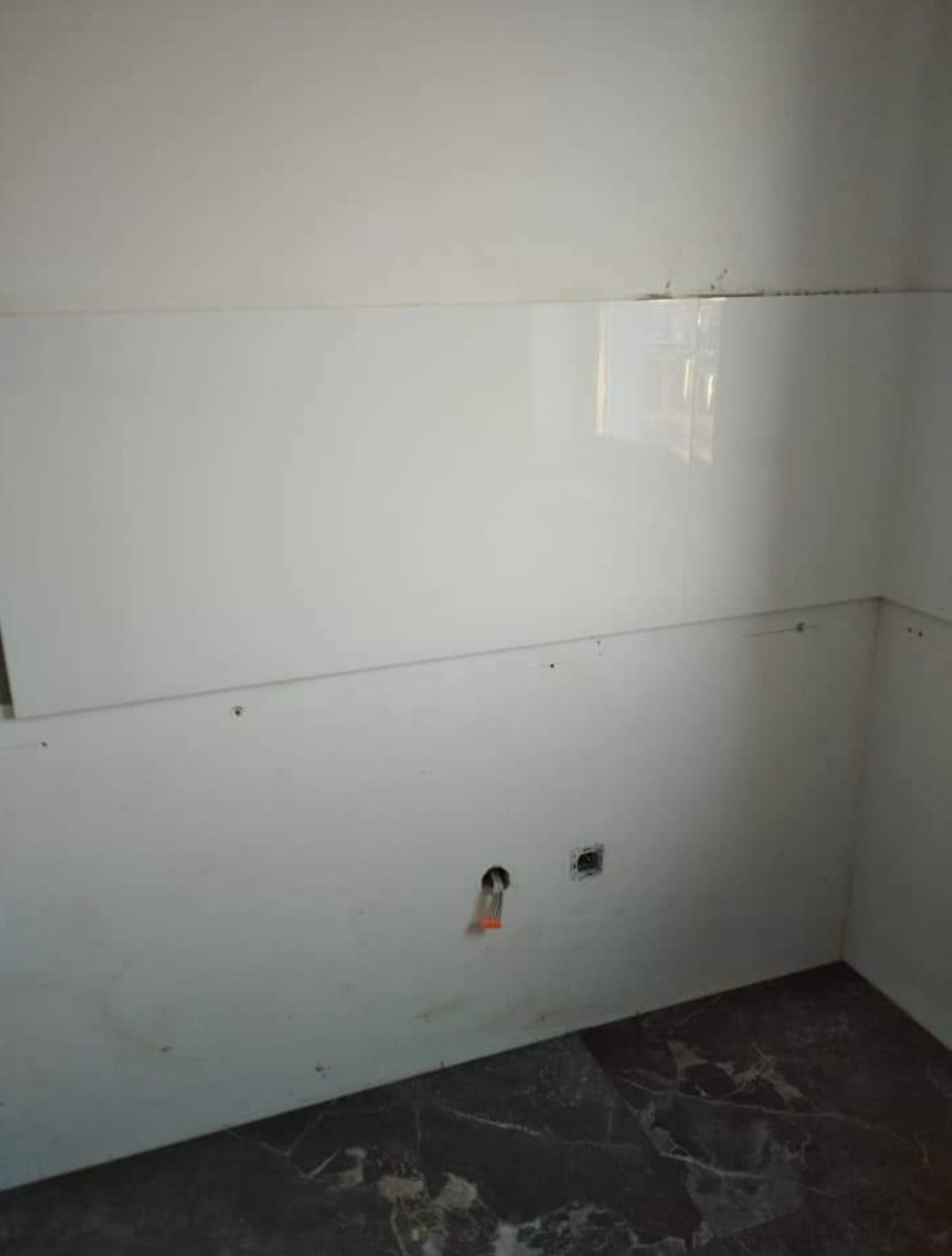 Fragment ściany w trakcie wykańczania z białymi, błyszczącymi panelami ściennymi, widoczne otwory na gniazdka i rury, marmurowa podłoga w odcieniach szarości.