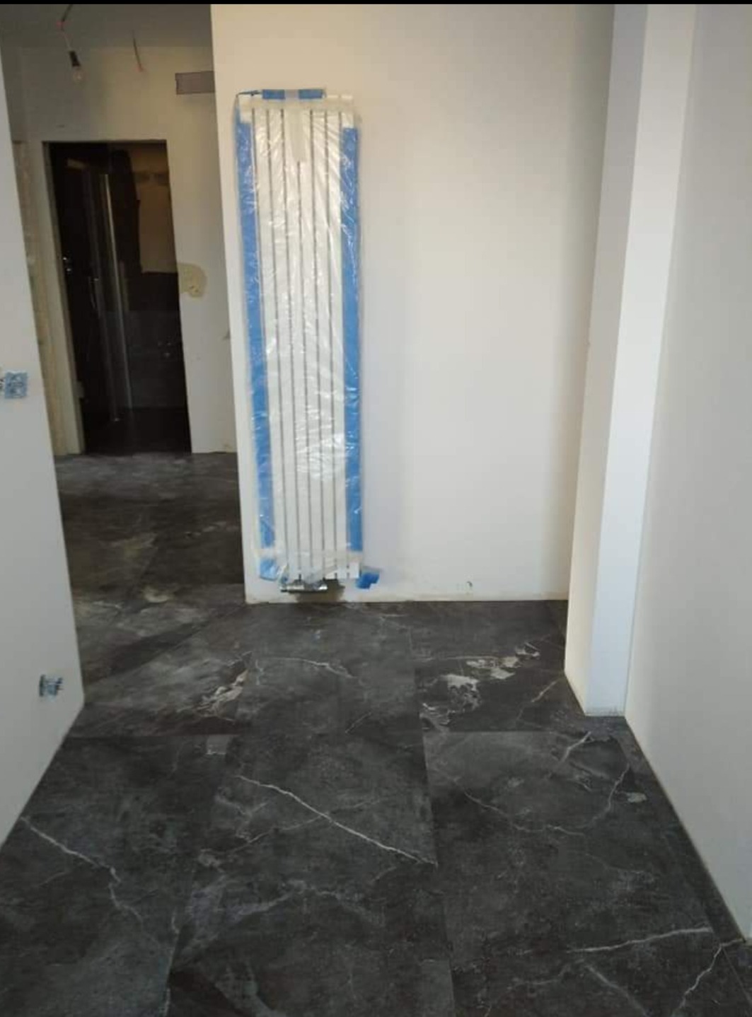 Nowe, ciemne płytki imitujące marmur na podłodze w jasnym pomieszczeniu, grzejnik zabezpieczony folią ochronną przed zabrudzeniem.
