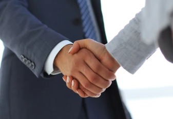 Uścisk dłoni dwóch osób, mężczyzny w granatowym garniturze i kobiety w szarej marynarce, symbolizujący zawarcie umowy lub partnerstwo biznesowe, na rozmytym, jasnym tle biurowym.