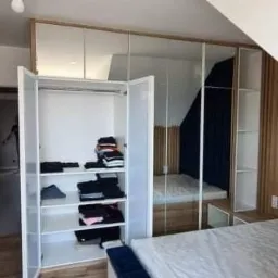 Przestronna szafa wnękowa z lustrzanymi drzwiami przesuwnymi i otwartą częścią z półkami, wykonana na wymiar w Rzeszowie. Wnętrze szafy zawiera ubrania ułożone na półkach oraz drążek na wieszaki.