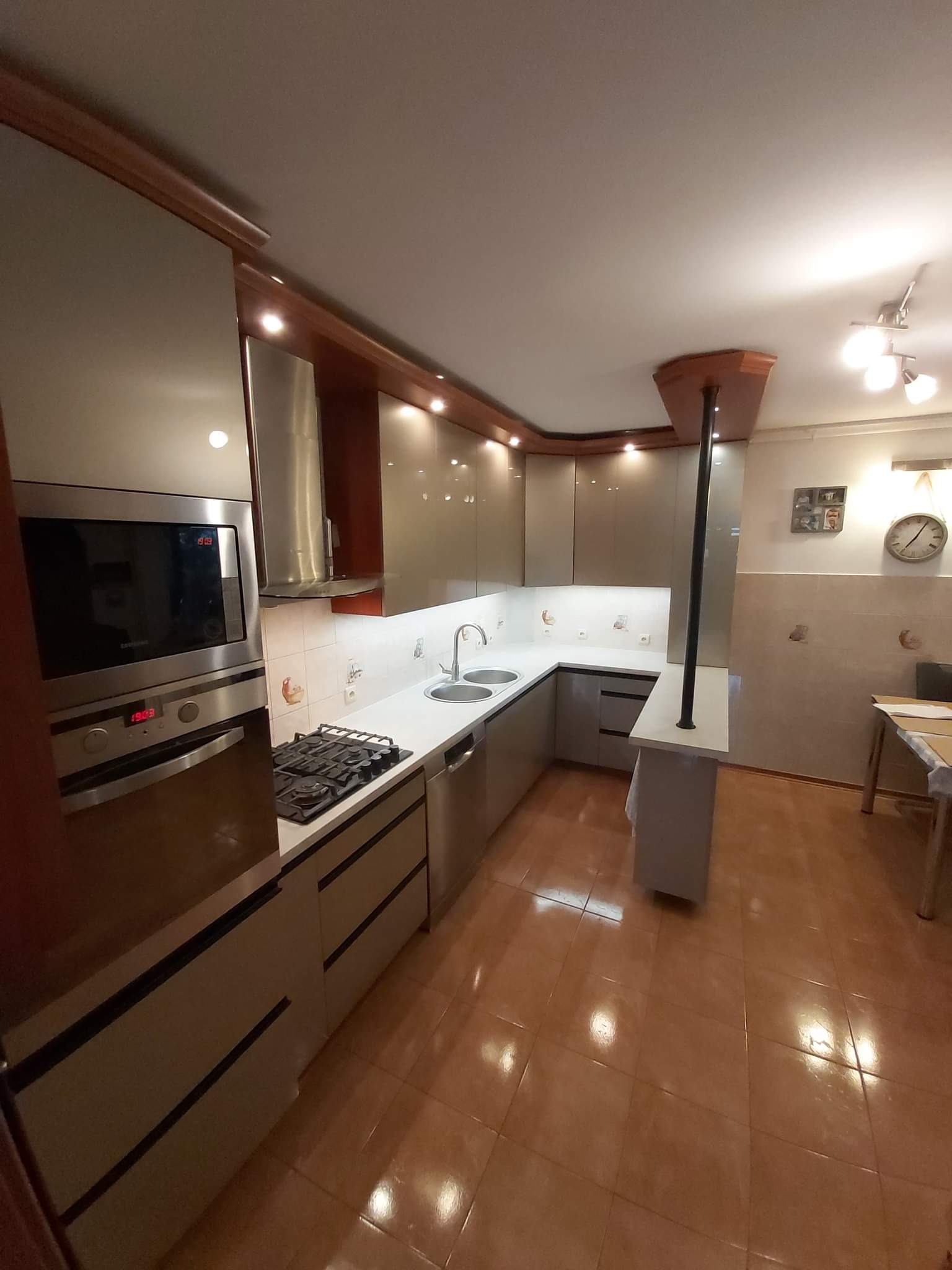 Jasna kuchnia z błyszczącymi frontami, oświetleniem podszafkowym i brązowymi akcentami, widok na płytę grzewczą, piekarnik i część blatu z dwukomorowym zlewem.