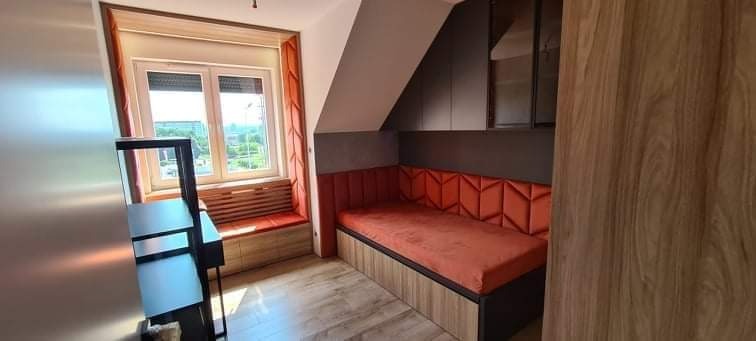 Pokój na poddaszu z zabudową: pomarańczowa tapicerowana wnęka z siedziskiem pod oknem i szafki nad łóżkiem.