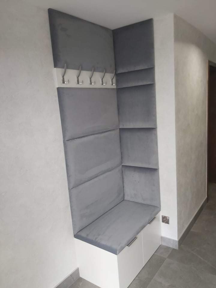 Szara, tapicerowana wnęka z siedziskiem i wieszakami, wbudowana w przedpokoju z białymi szafkami na dole.