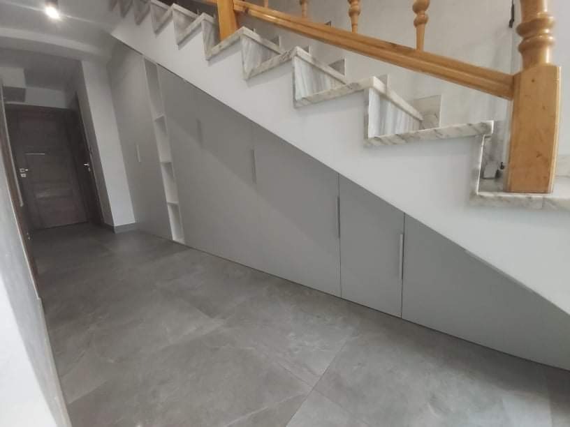 Szare szafki z minimalistycznymi uchwytami wkomponowane pod marmurowe schody z drewnianą balustradą, szara podłoga z płytek.