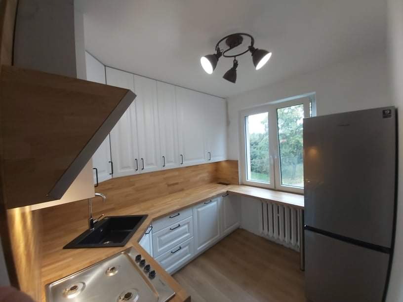 Jasna kuchnia z drewnianym blatem, białymi szafkami z czarnymi uchwytami i czarnym zlewozmywakiem, okap z drewnianym wykończeniem, widok na okno i grzejnik.