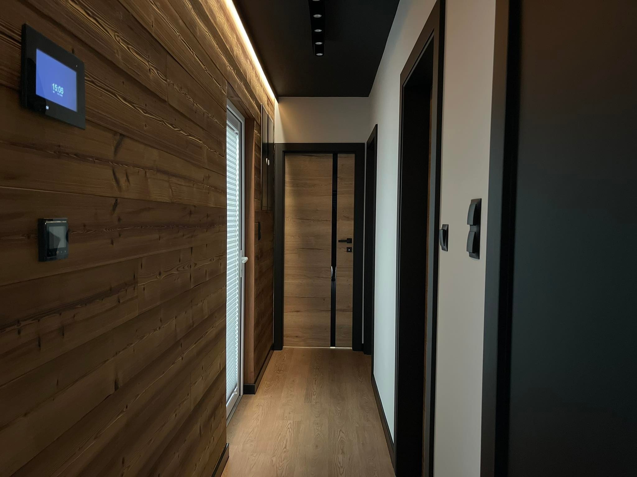 Nowoczesny korytarz z boazerią z ciemnego drewna, czarnym sufitem z oświetleniem punktowym i drewnianymi drzwiami z czarnymi framugami, widoczny panel sterowania inteligentnego domu na ścianie.