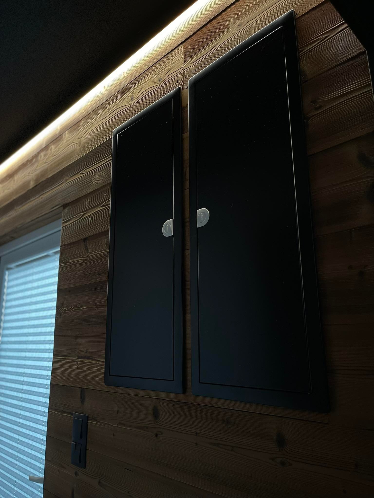 Dwa czarne panele rewizyjne z chromowanymi zamkami, zamontowane na ścianie wyłożonej drewnianą boazerią z podświetleniem LED.