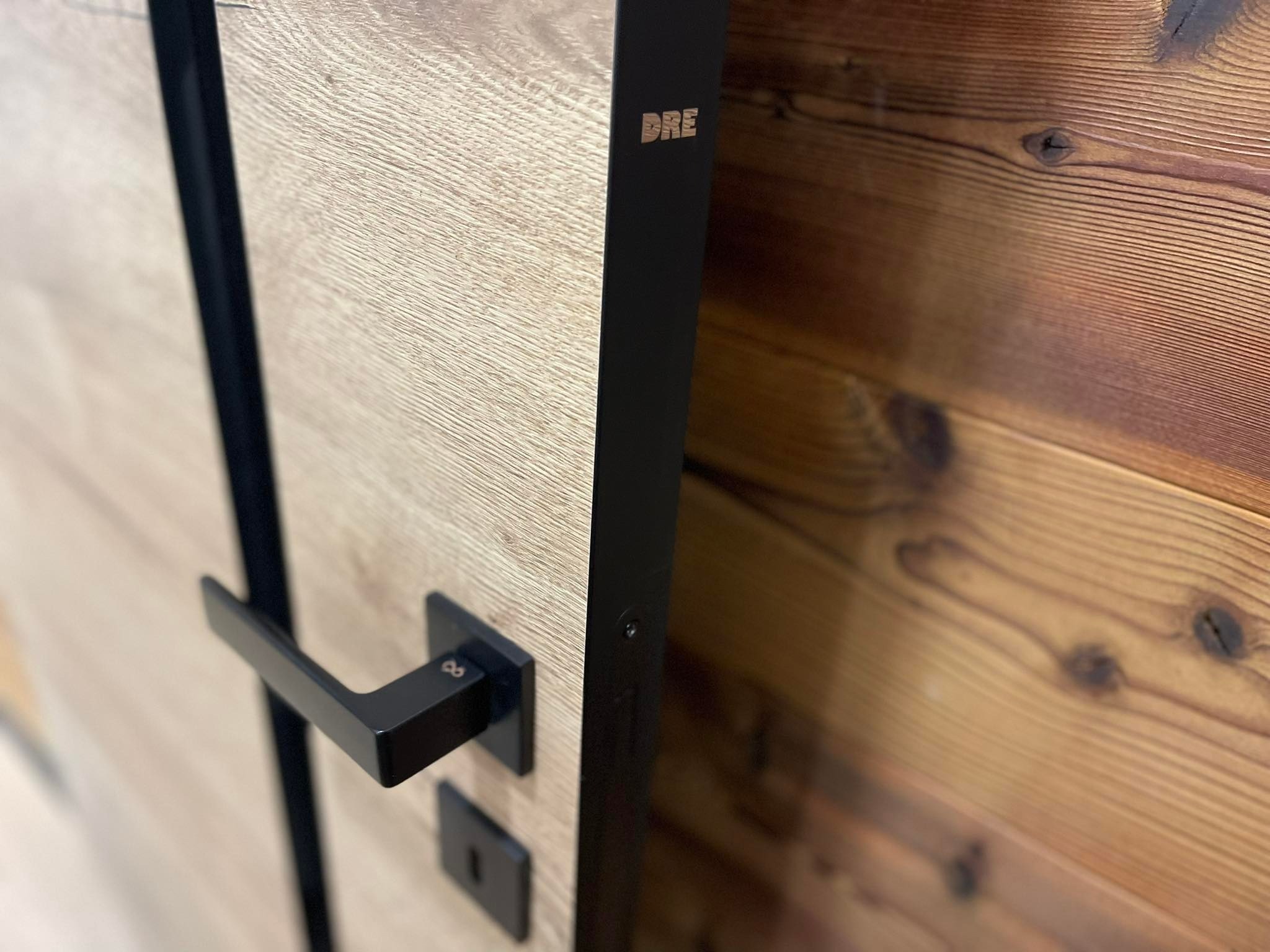 Fragment drewnianej ściany z widocznymi słojami drewna i zamontowane drzwi z czarną klamką oraz czarną futryną z widocznym logo DRE w górnej części.