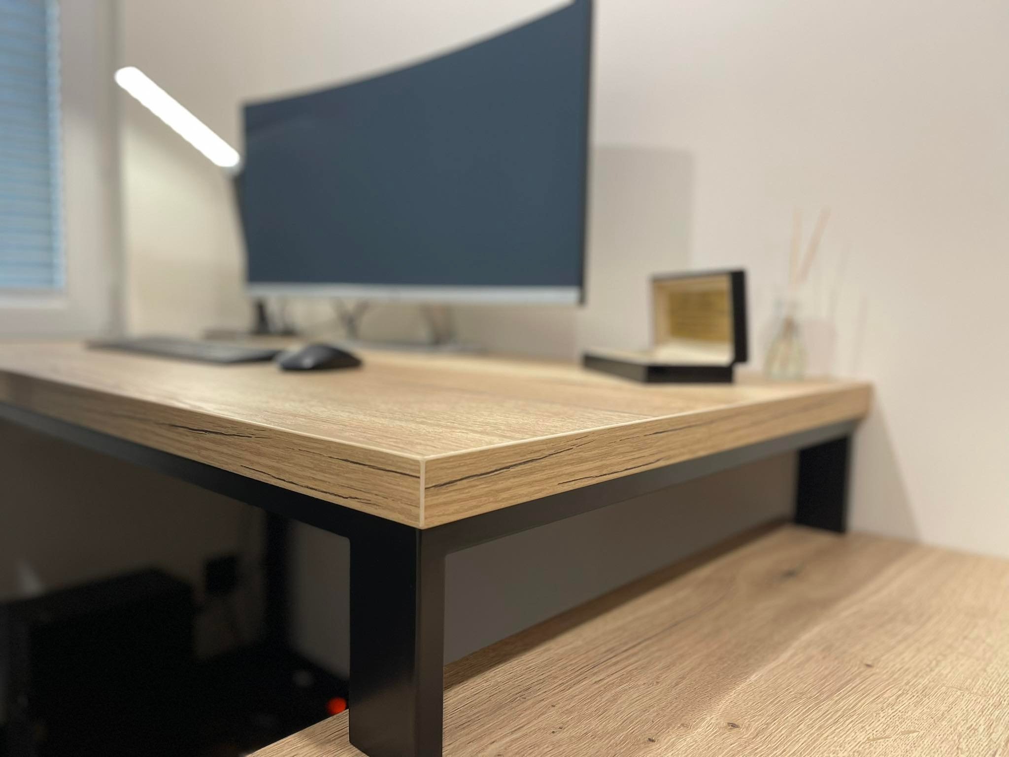 Biurko z blatem imitującym jasne drewno, czarne metalowe nogi, zakrzywiony monitor, klawiatura i mysz na blacie, minimalistyczna lampka i ozdobne pudełko w tle.