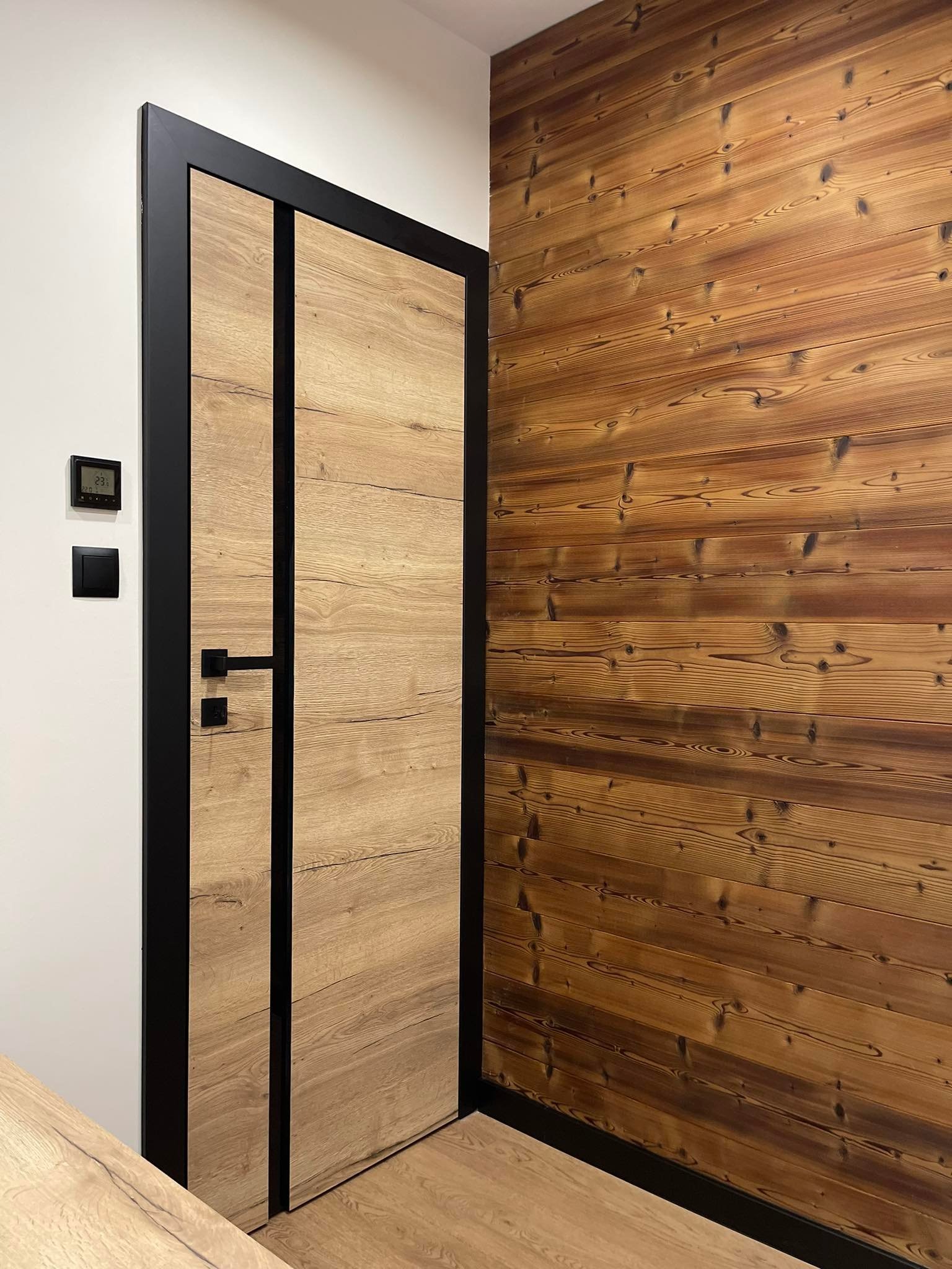 Drewniana ściana z boazerii i drzwi z czarną ramą, widoczny termostat na ścianie.