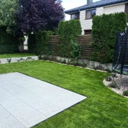 Nowoczesny ogród z jasną, kwadratową płytą tarasową, świeżo ułożoną zieloną trawą z rolki, ozdobnymi kamieniami i trampoliną w tle.