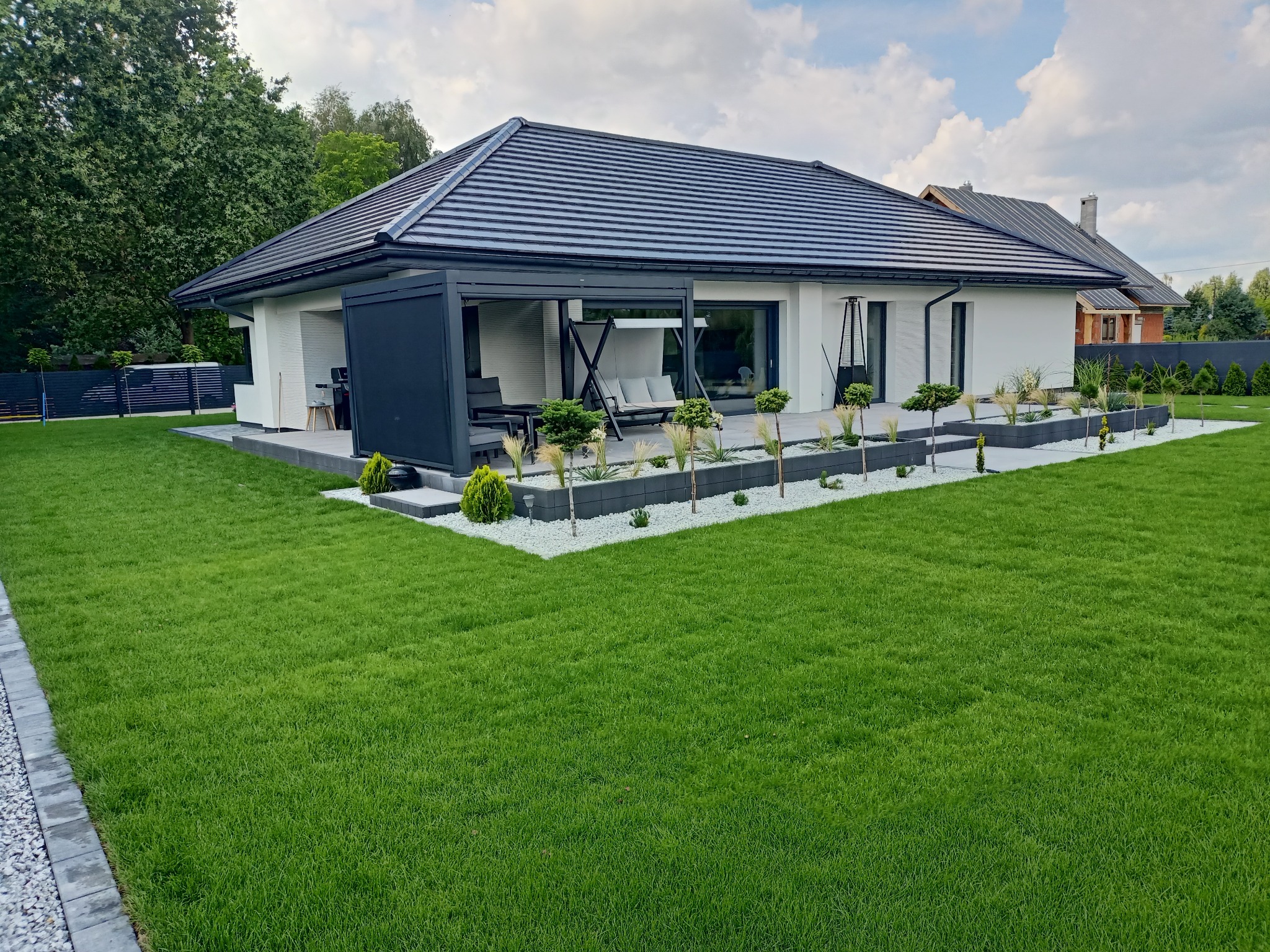 Nowoczesny dom z zadbanym trawnikiem i minimalistycznym ogrodem. Widoczny taras z pergolą i meblami ogrodowymi. Białe kamienie dekoracyjne i niskie drzewka.