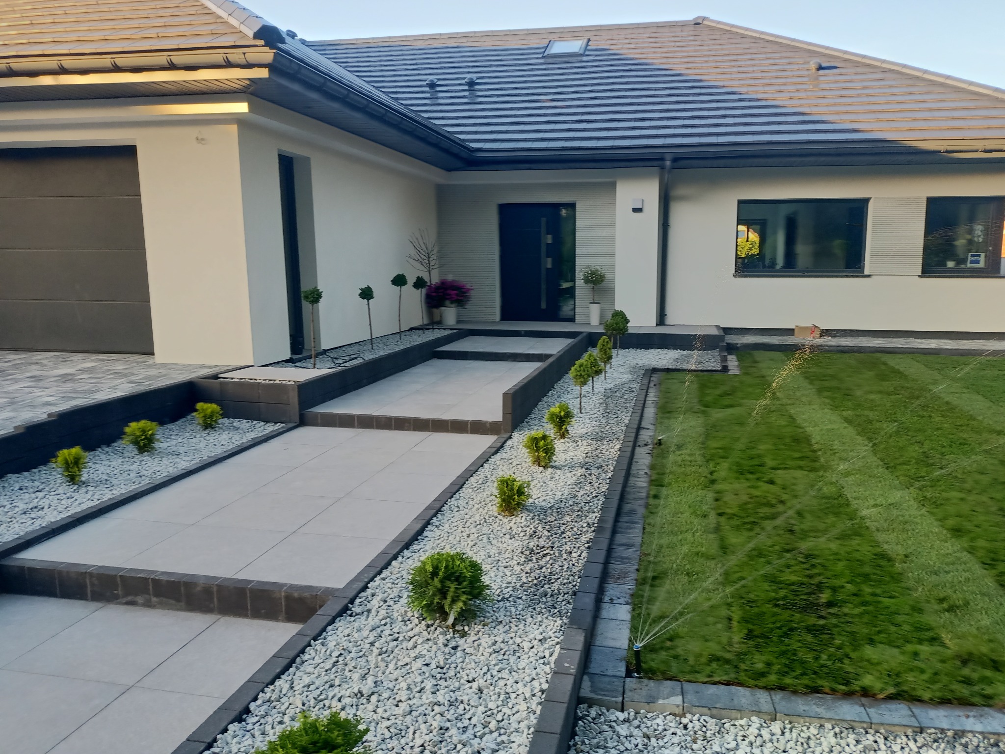 Nowoczesny ogród z równo przystrzyżonym trawnikiem, systemem nawadniania, ścieżką z jasnych płyt i białych kamieni, prowadzącą do domu z szarą dachówką.