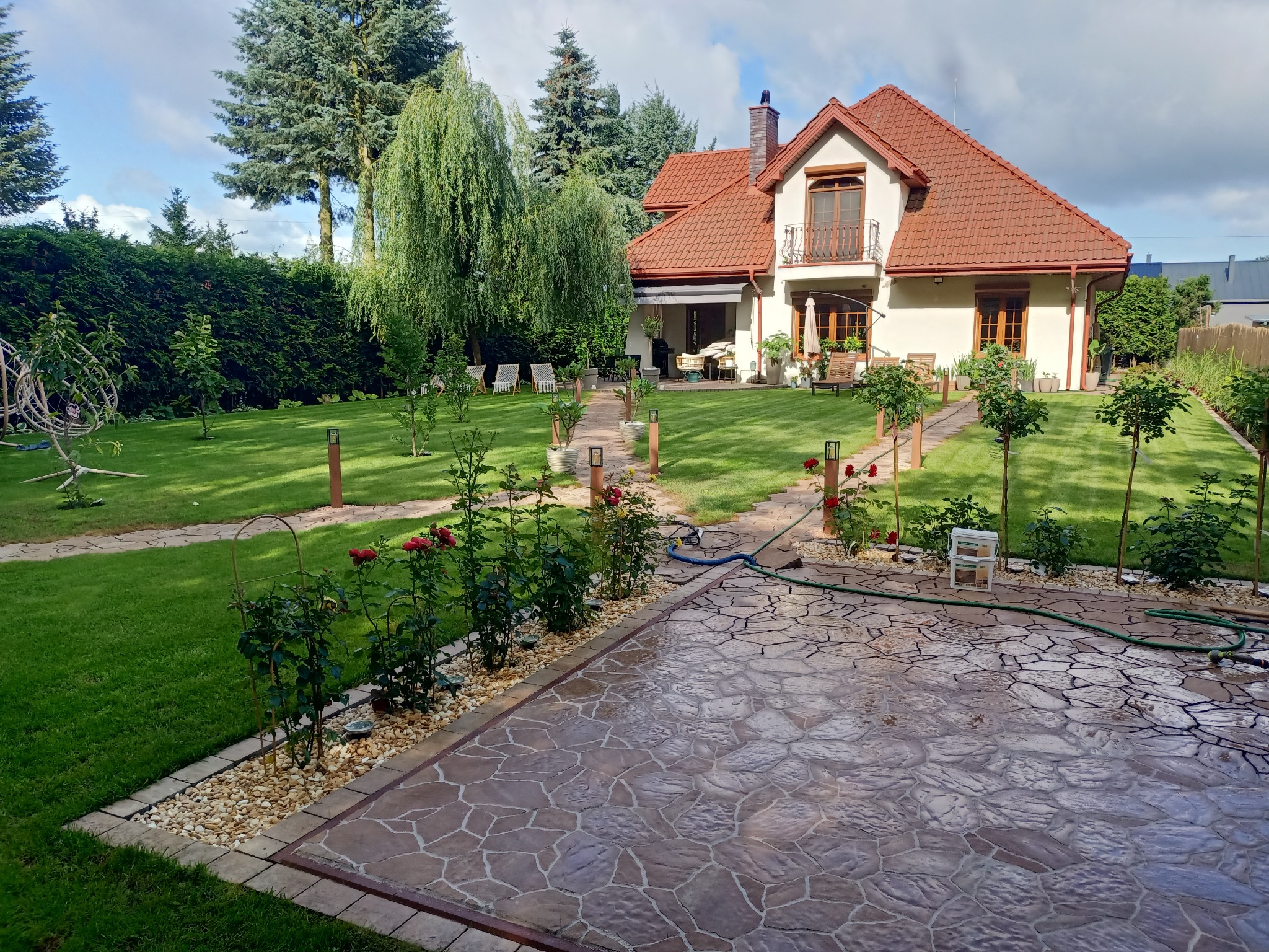 Rozległy ogród z brukowanym tarasem i ścieżką, rabatami róż, oświetleniem i zadbanym trawnikiem, prowadzący do domu z czerwonym dachem i balkonem.
