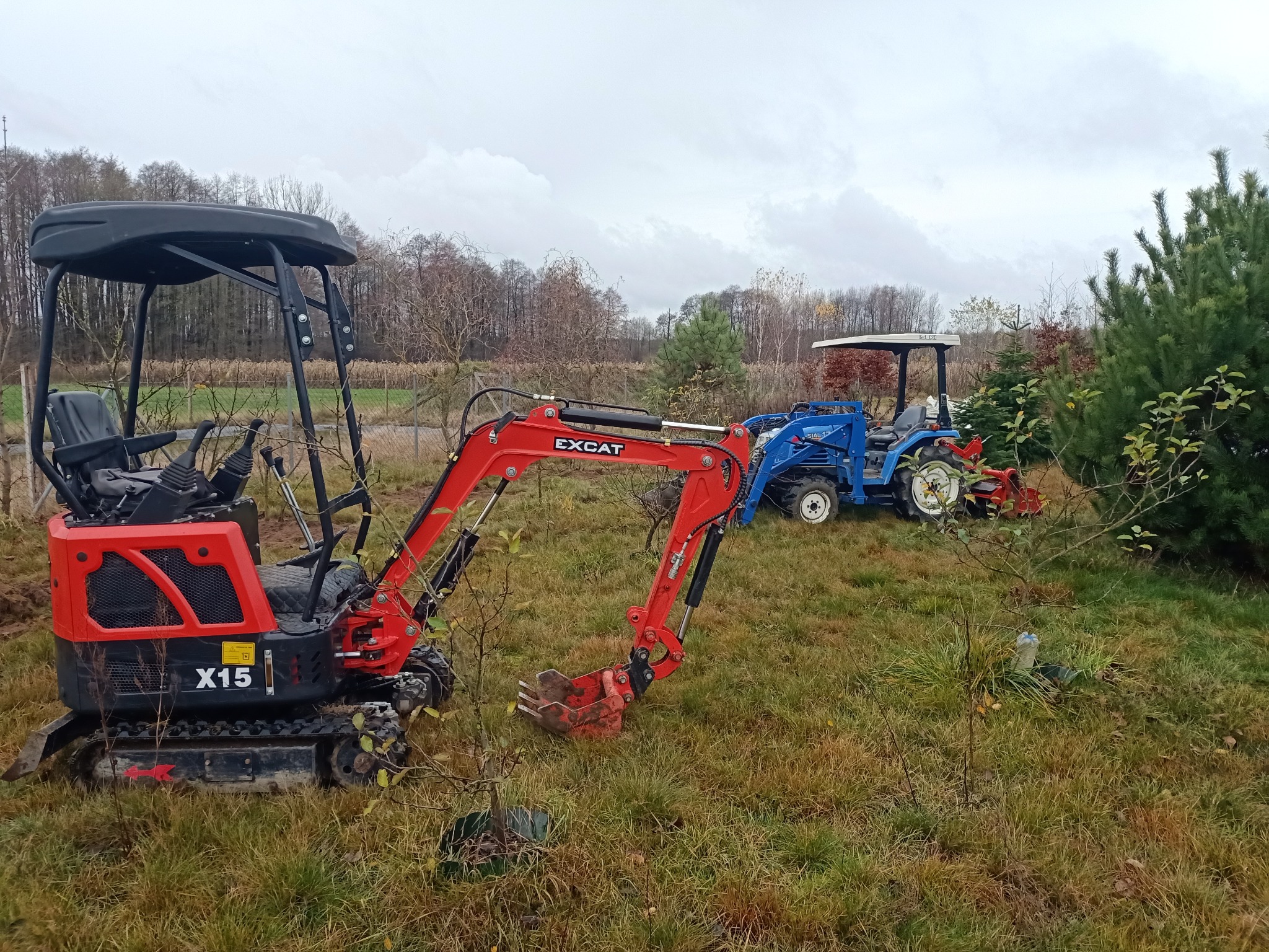 Czerwony minikoparka EXCAT X15 na trawiastym terenie, obok niebieski traktor z białym dachem, w tle drzewa pod pochmurnym niebem. Sprzęt gotowy do pracy.