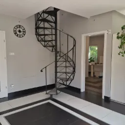 Wnętrze biura z metalowymi schodami spiralnymi, zegarem na ścianie i rośliną doniczkową na szafie.