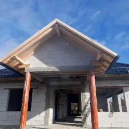 Budowa domu jednorodzinnego z widocznym drewnianym szkieletem dachu i częściowo pokrytym dachem z blachy, widoczne pustaki i pomarańczowe rury spustowe.