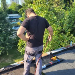 Dekarz z palnikiem na dachu, z tyłu widok na zieleń i budynki mieszkalne, w tle butelka wody i narzędzia.